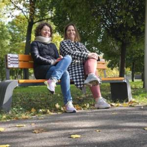 Dwie osoby siedzą na pomarańczowej ławce z tabliczką numerów pomocowych w parku; obok alejka z jesiennymi liśćmi.