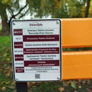 Zbliżenie tabliczki na ławce z numerami telefonów zaufania; krople deszczu na lakierze