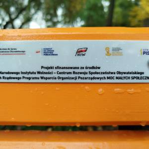 Zbliżenie tabliczki informującej o finansowaniu projektu przez NIW i program MOC MAŁYCH SPOŁECZNOŚCI; krople deszczu.