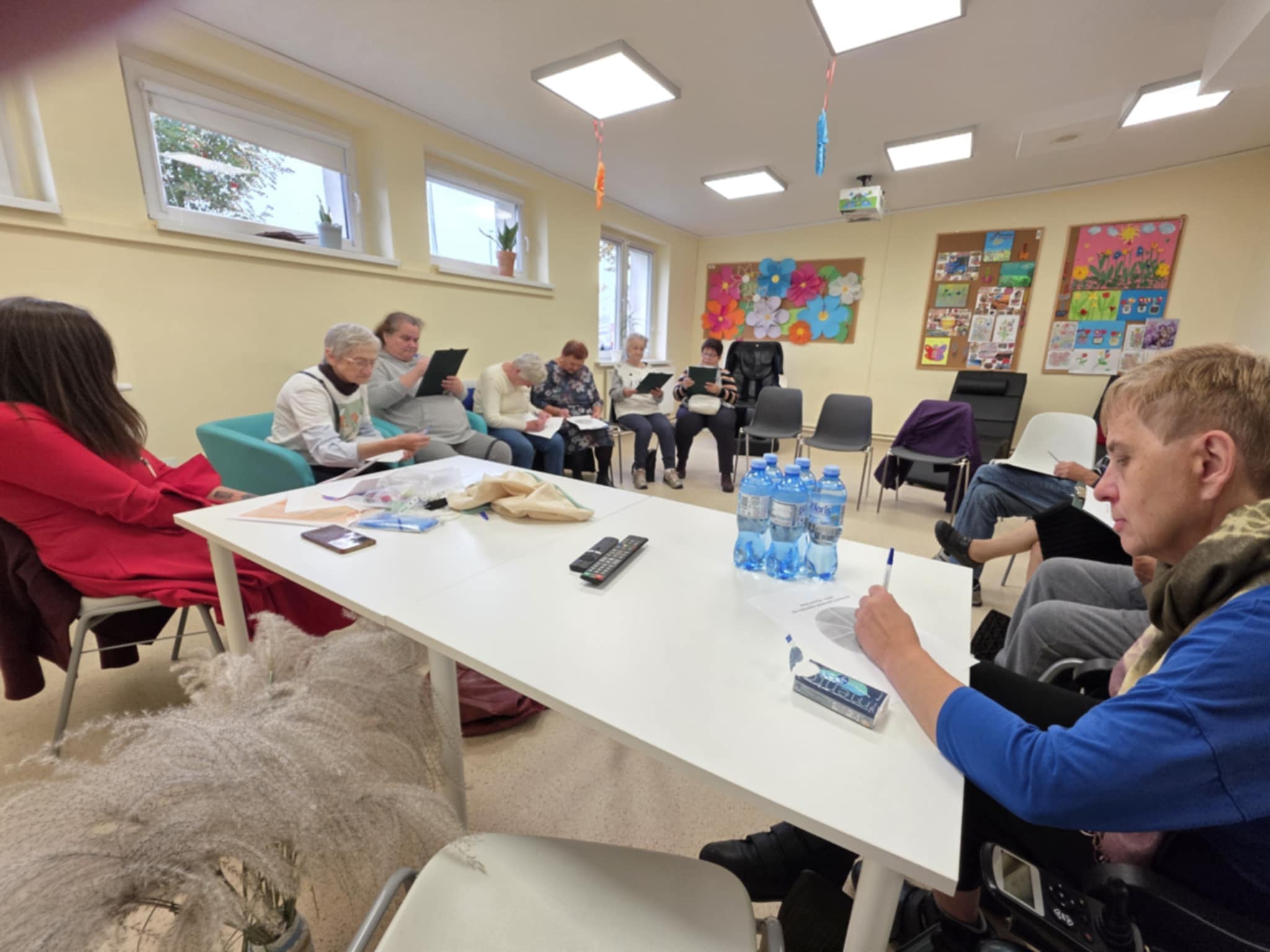 Grupa uczestników w sali podczas warsztatów; siedzą w kręgu, pracują na kartach, na stole woda i materiały.