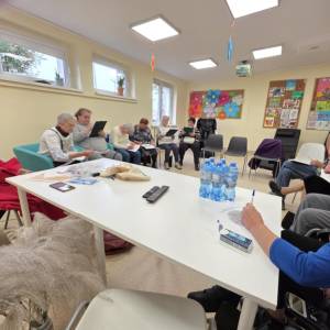 Grupa uczestników w sali podczas warsztatów; siedzą w kręgu, pracują na kartach, na stole woda i materiały.