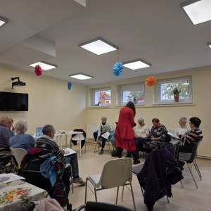 Warsztaty w sali ŚDS; prowadząca w czerwieni pomaga przy stoliku, uczestnicy czytają materiały.