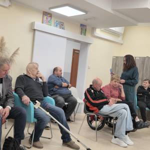Spotkanie w ŚDS; uczestnicy siedzą w kręgu, prowadząca pokazuje materiał dwóm osobom, w tle kule ortopedyczne.