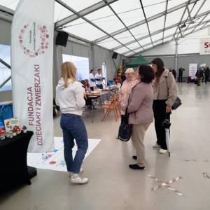 Hala wydarzeń. Stoisko „Fundacja Dzieciaki i Zwierzaki”, animatorka rozmawia z trzema kobietami. W tle inne stoiska i baner „Swarzędz”.