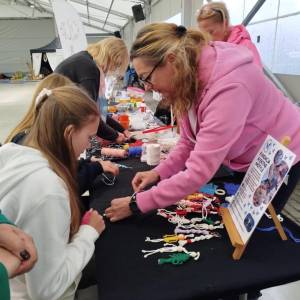 Warsztaty makramy. Instruktorka w różowej bluzie pomaga dzieciom i dorosłym robić bransoletki na czarnym stole z kolorowymi sznurkami.