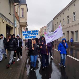 Marsz uliczny. Grupa młodzieży i dorosłych niesie tablicę „Każdy inny, wszyscy równi” oraz flagę „uMocnieni w kryzysie”.