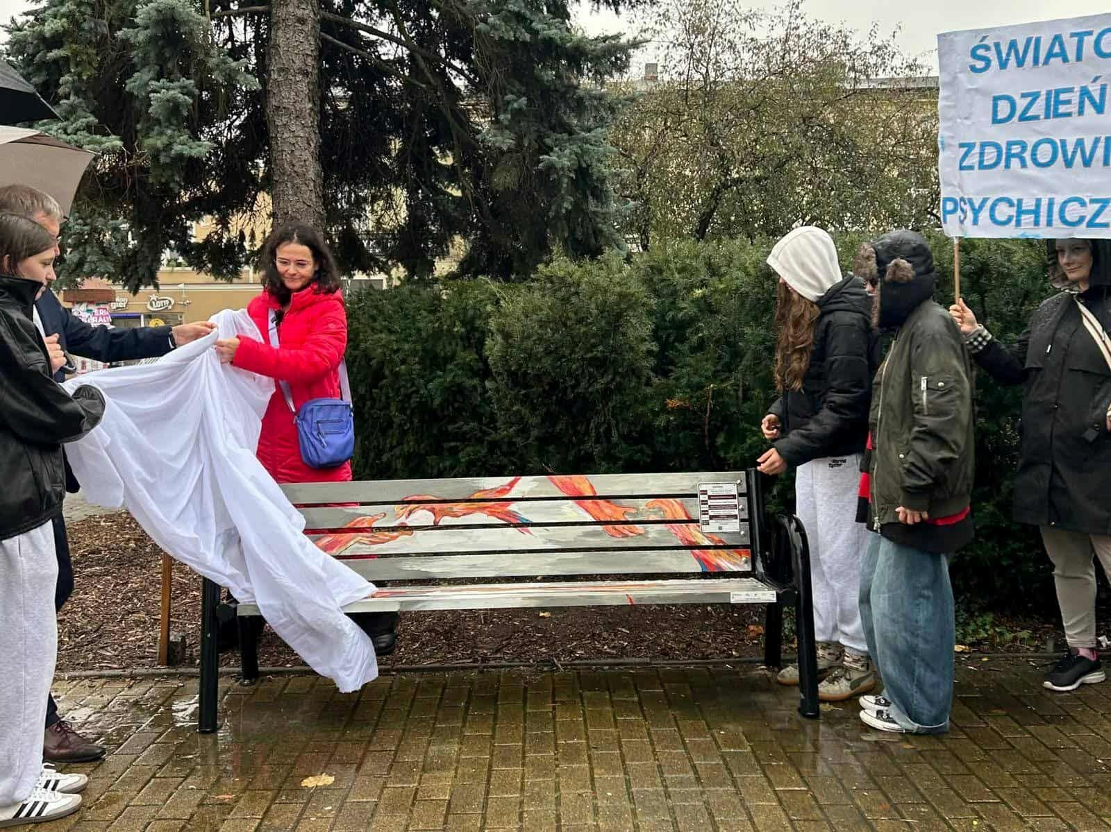 Grupa osób odsłania kolorową „Ławkę Dobrych Słów” w parku podczas Światowego Dnia Zdrowia Psychicznego; na mokrym chodniku widać ławkę z ilustracją i tabliczką informacyjną.