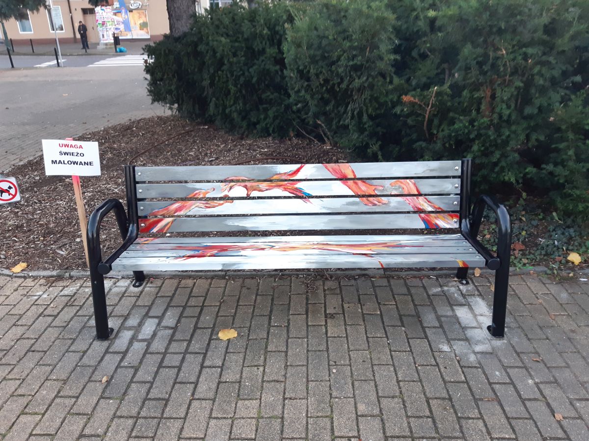 Ławka z artystycznym malunkiem w odcieniach czerwieni i szarości, z tabliczką ‘świeżo malowane’ obok.