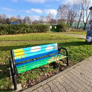 Ławka z malunkiem słońca, chmur i zieleni, stojąca na trawniku przy chodniku i słupie ogłoszeniowym.