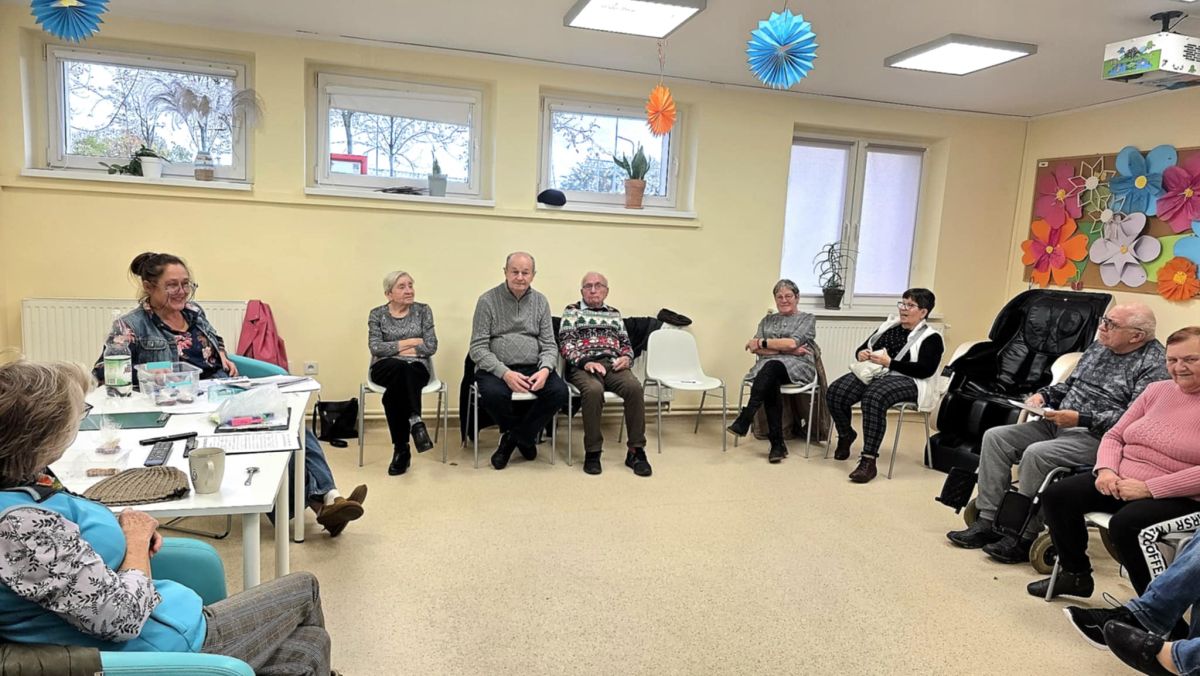 Grupa seniorów siedzi w kręgu na zajęciach w jasnej sali, rozmawiają i uczestniczą w spotkaniu integracyjnym
