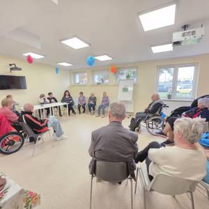 Seniorzy siedzą w dużym kręgu podczas zajęć w świetlicy; w sali widać dekoracje i osoby na wózkach