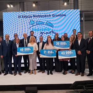 Uroczystość Niebieskich Grantów — laureaci i przedstawiciele organizatorów stoją razem z czekami, widoczny baner III Edycji Niebieskich Grantów.