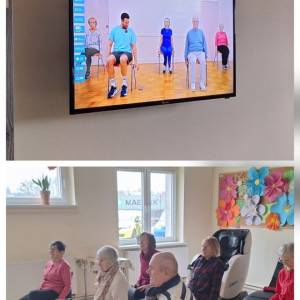 Seniorzy siedzą w sali na krzesłach, uczestniczą w ćwiczeniach rehabilitacyjnych oglądając instrukcje na telewizorze, wspólne zajęcia w DDP