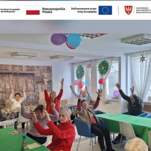 Seniorzy siedzą przy stolikach i wspólnie unoszą ręce podczas zajęć gimnastycznych w sali Dziennego Domu Pomocy, ćwiczenia grupowe