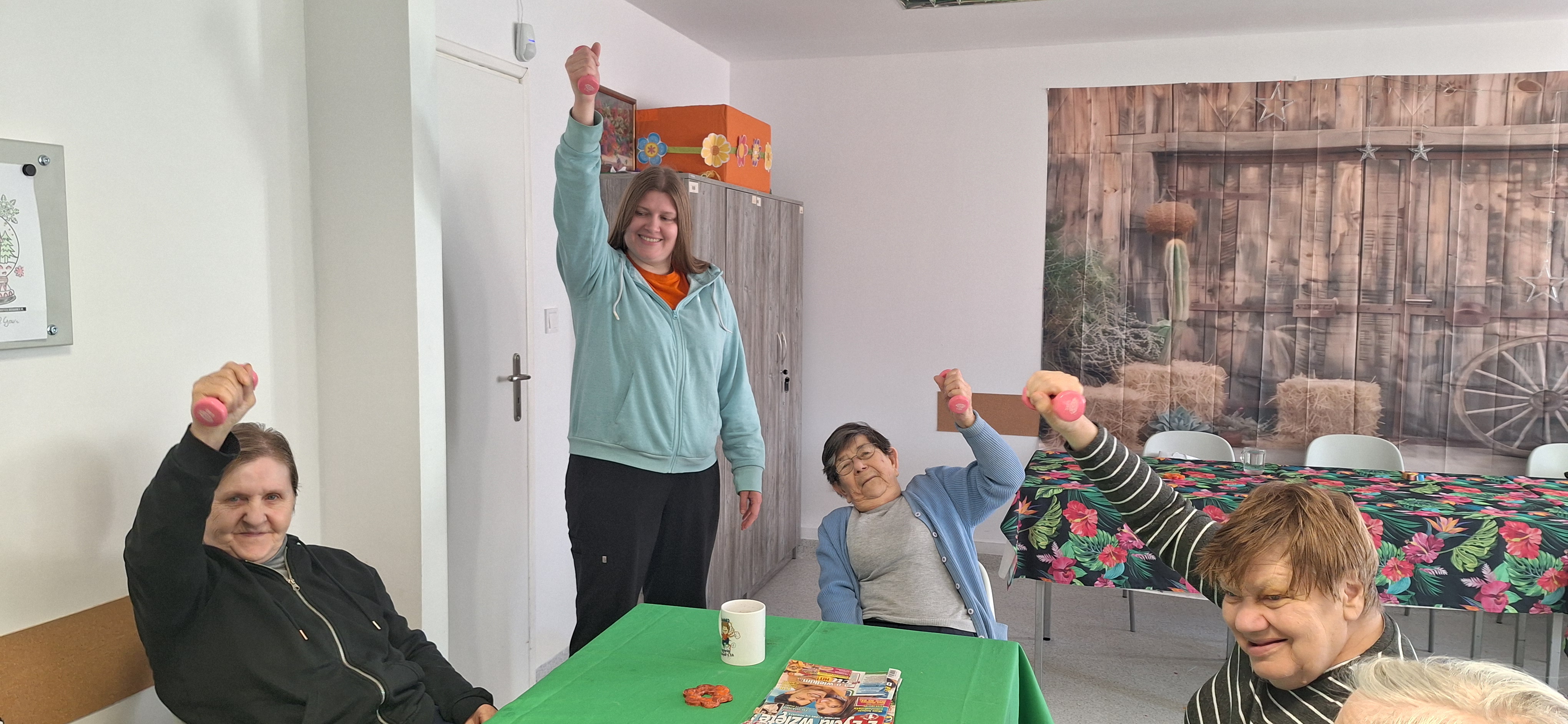 Seniorzy siedzą przy stolikach i unoszą ręce z małymi hantelkami podczas ćwiczeń rehabilitacyjnych w Dziennym Domu Pomocy.