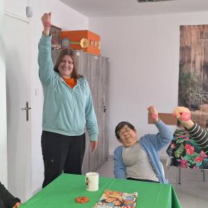 Seniorzy siedzą przy stolikach i unoszą ręce z małymi hantelkami podczas ćwiczeń rehabilitacyjnych w Dziennym Domu Pomocy.