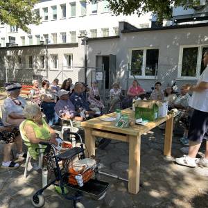 Grupa osób słucha wykładowcy w ogrodzie społecznym Centrum Usług Społecznych w Swarzędzu. Na stole przed wykładowcą widać preparaty ogrodnicze takie jak nawóz do roślin. W tle stoi budynek Centrum Usług Społecznych w Swarzędzu.