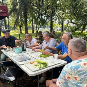Grupa seniorów siedząca przy połączonych stołach w plenerze podczas warsztatów kulinarnych 'Ogród Pokoleń'. Uczestnicy obierają i kroją świeże warzywa oraz owoce, przygotowując m.in. ogórki na tacach. W tle znajduje się bordowy namiot i parkowa zieleń.