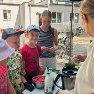 Dzieci przy stole kuchennym obserwują z bliska przygotowywanie orzeźwiającego koktajlu podczas plenerowych warsztatów 'Ogród Pokoleń'. Instruktorka po prawej stronie wrzuca składniki do robota kuchennego, a przed nią stoi miska pokrojonego arbuza i butelki z maślanką. Uczestnicy z zainteresowaniem śledzą pokazy kulinarne.