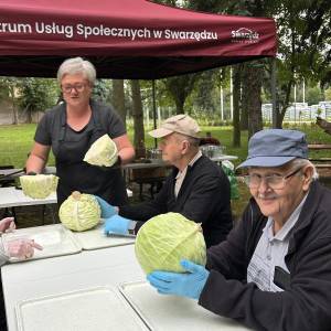 Uśmiechnięty senior w niebieskiej czapce z daszkiem i rękawiczkach ochronnych prezentuje dużą główkę kapusty podczas warsztatów przetwórczych w Swarzędzu. Obok przy stole instruktorka rozdaje kolejne porcje warzyw uczestnikom siedzącym pod namiotem Centrum Usług Społecznych. Spotkanie odbywa się na świeżym powietrzu w Ogrodzie Społecznym.