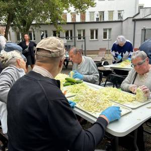 Opis alternatywny:

Seniorki i seniorzy siedzą przy stole plenerowym i wspólnie szatkują główki białej kapusty na dużych białych tacach. Uczestnicy w rękawiczkach ochronnych kroją warzywa, przygotowując je do kiszenia w ramach warsztatów w Ogrodzie Społecznym. W tle widać kolejne stanowiska z pracującymi uczestnikami oraz budynek i ogrodowe drzewa.