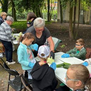 Pani Ania instruuje grupę dzieci z placówki 'Nasza Dziupla' podczas przygotowywania warzyw na warsztatach w Ogrodzie Społecznym w Swarzędzu. Dzieci w rękawiczkach ochronnych siedzą wokół stołu, na którym leżą świeże ogórki oraz szatkowana kapusta. W tle, pod drzewami, widać seniorki pracujące przy sąsiednim stole plenerowym.