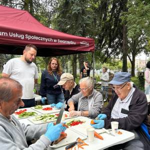 Seniorki i seniorzy przygotowują warzywa, siedząc przy stole pod czerwonym namiotem z napisem 
