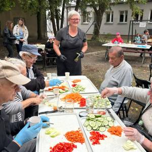 Seniorki i seniorzy siedzą przy stole w Ogrodzie Społecznym w Swarzędzu, przygotowując pokrojone w plasterki i kostkę warzywa: ogórki, marchewkę i paprykę do sałatki w słoikach. Nad przebiegiem prac czuwa stojąca pośrodku instruktorka w czarnym fartuchu i okularach. W tle, przy kolejnych stołach na świeżym powietrzu, widać dzieci i pozostałych uczestników międzypokoleniowych warsztatów.