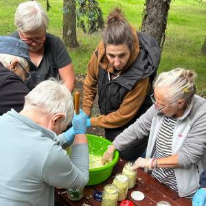 Uczestnicy warsztatów przetwórczych w Ogrodzie Społecznym w Swarzędzu wspólnie przygotowują kiszoną kapustę przy drewnianym stole na świeżym powietrzu. Seniorzy i instruktorka ubijają szatkowaną kapustę w zielonej misce i napełniają nią szklane słoiki, pracując w rękawiczkach ochronnych. Spotkanie odbywa się w otoczeniu ogrodowej zieleni i drzew.