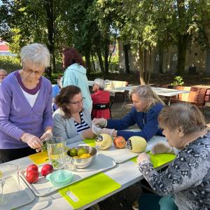 Seniorki siedzą i stoją przy plenerowym stole w Ogrodzie Społecznym, przygotowując owoce uratowane z Banku Żywności na sałatkę i orzeźwiający kompot. Na tacach i w miskach przed uczestniczkami w rękawiczkach ochronnych leżą melony, granaty, nektarynki, cytrusy oraz świeża mięta. Warsztaty kulinarne zero waste odbywają się na świeżym powietrzu w cieniu ogrodowych drzew.