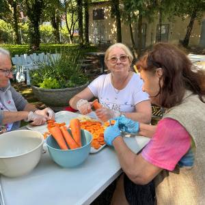 Uczestniczki warsztatów kulinarnych zero waste siedzą przy stole w Ogrodzie Społecznym, wspólnie obierając i krojąc marchewkę na zupę krem. Kobiety w rękawiczkach ochronnych napełniają miski oraz garnki świeżymi warzywami pozyskanymi z Banku Żywności. W tle widać słoneczną, zieloną przestrzeń ogrodu z roślinnością i drzewami.