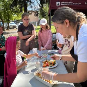 Pani Monika z inicjatywy 'Siedzę w Garach' nakłada pieczone owoce i warzywa do papierowych kubeczków dla dzieci podczas warsztatów kulinarnych zero waste w Ogrodzie Społecznym. Najmłodsi uczestnicy z zaciekawieniem czekają przy stole na degustację przygotowanych potraw. W tle widoczny jest bordowy namiot Centrum Usług Społecznych w Swarzędzu oraz słoneczny ogród.