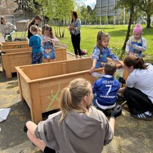 Dzieci i młodzież wspólnie ozdabiają farbami duże drewniane skrzynie uprawne ustawione na świeżym powietrzu w Ogrodzie Społecznym. Uczestnicy w rękawiczkach ochronnych malują zielone wzory na deskach, a w tle widać opiekunów oraz słoneczny, zielony teren przygotowywany pod ogród w ramach projektu „Ogród Pokoleń”.